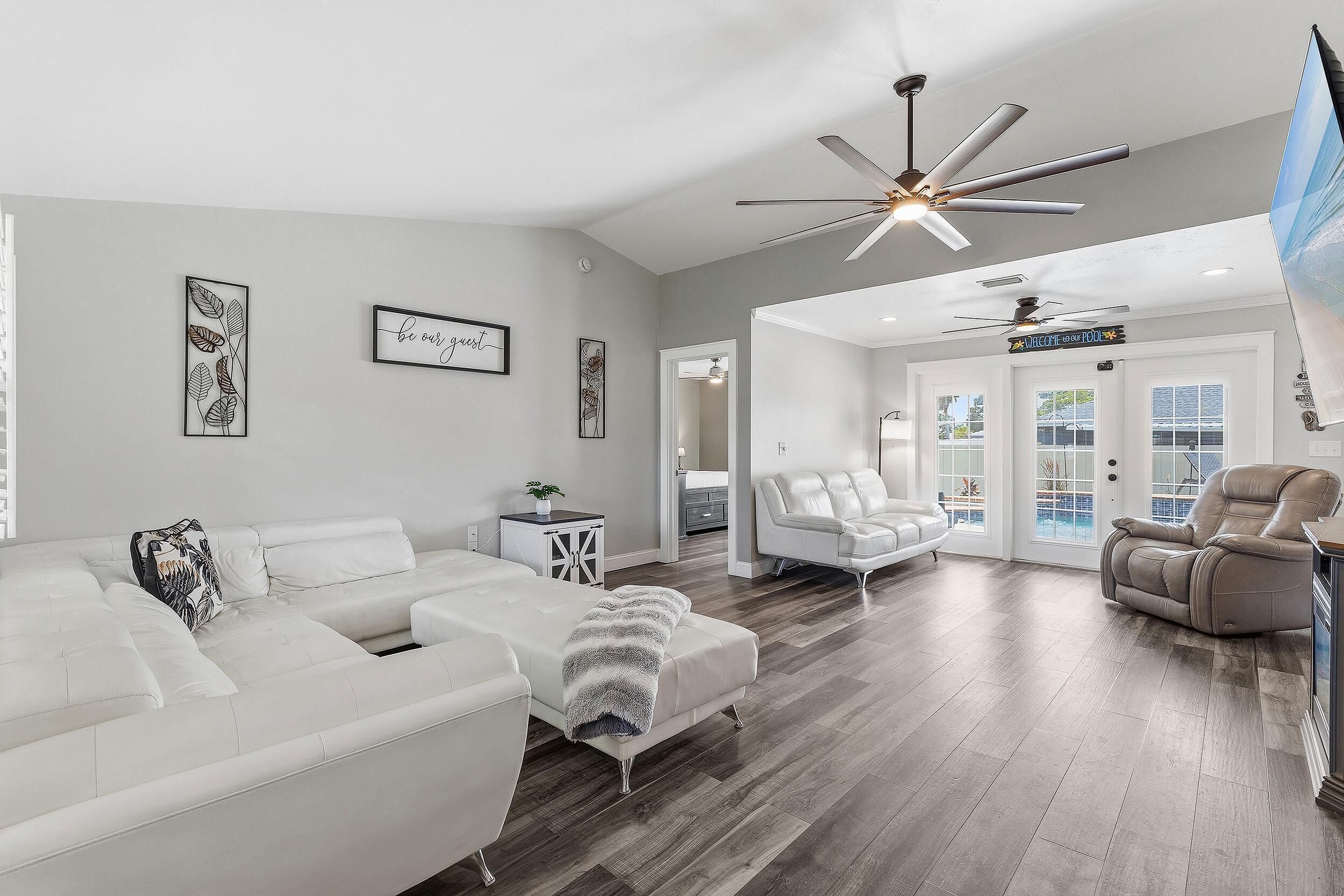 A modern living room with a white sofa, a recliner, a ceiling fan, and a sign reading 'Welcome & Pool'.