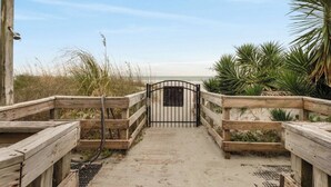 En træpromenade fører hen til en strand med en metalport, omgivet af sand og vegetation.