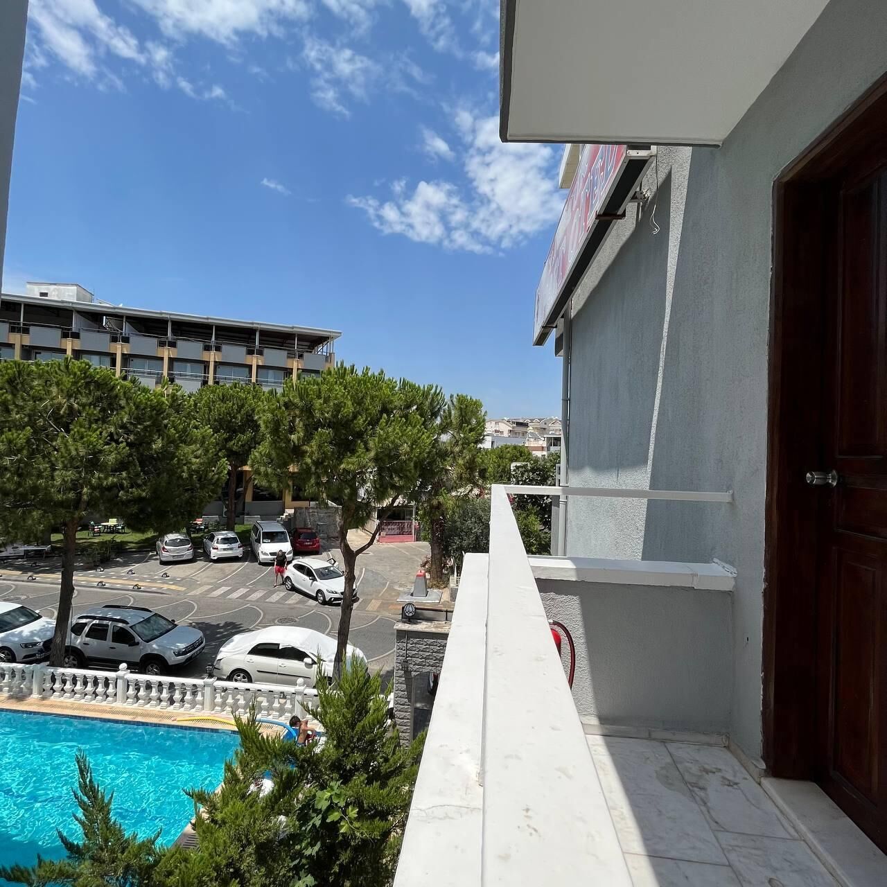 A balcony with a view of a swimming pool, parked cars, and a building in the background.