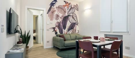A modern living room with a green sofa, a dining table with chairs, a flat-screen TV, and a wall mural.