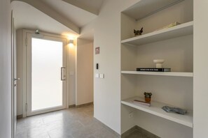A hallway with a door leading to a bright room, a shelf with books and decorative items, and a sign indicating a waiting area.