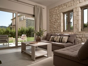 A modern living room with a stone wall, a sofa set, a coffee table, and a view of a stone building and greenery outside. - Avesta Private Villas Iokasti (Lefkada)