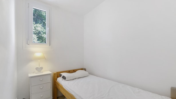 A small, single-bed room with a wooden headboard, a bedside table with drawers, a lamp, and a window with a view of trees.
