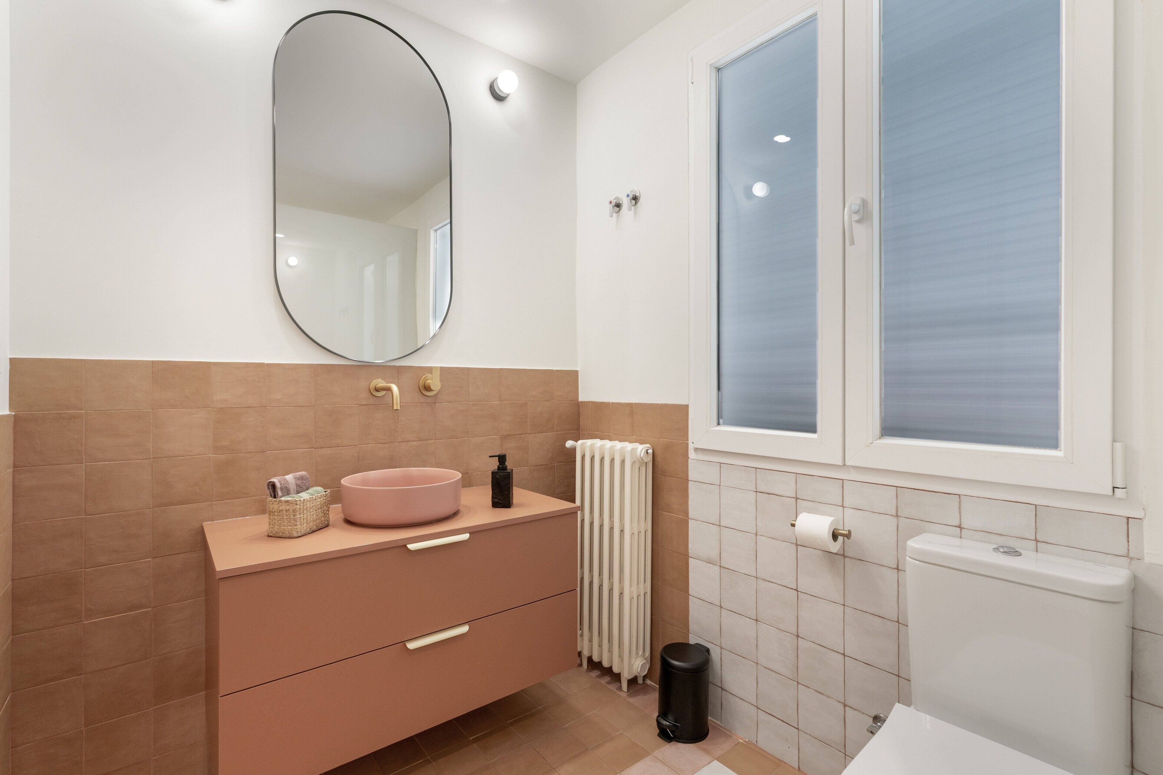 Ein Badezimmer mit einem rosafarbenen Waschtisch, einem runden Spiegel, einer Toilette und einem Fenster mit Milchglas.