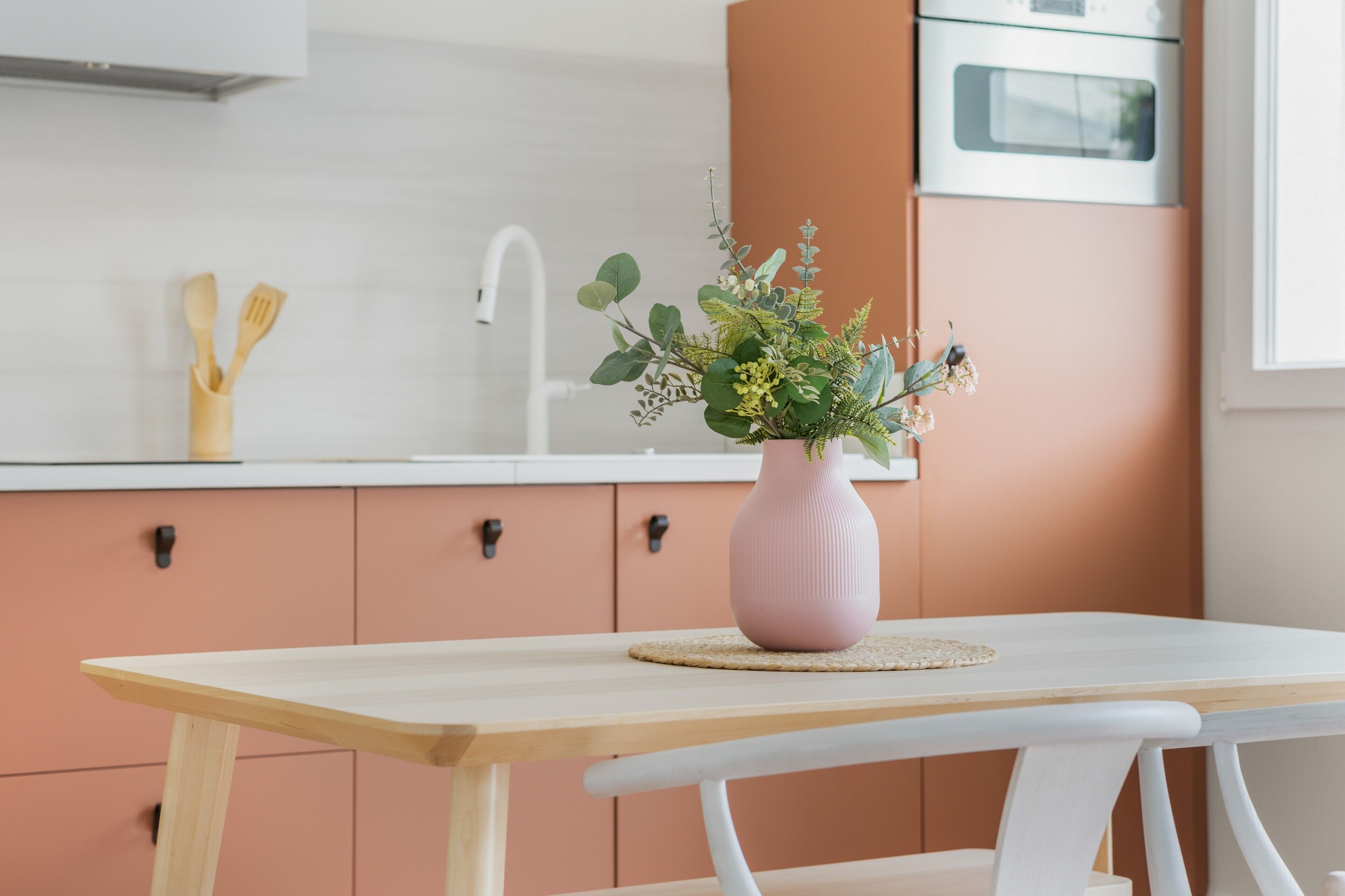 Een keuken met een houten tafel, een roze vaas met bloemen en een witte gootsteen.