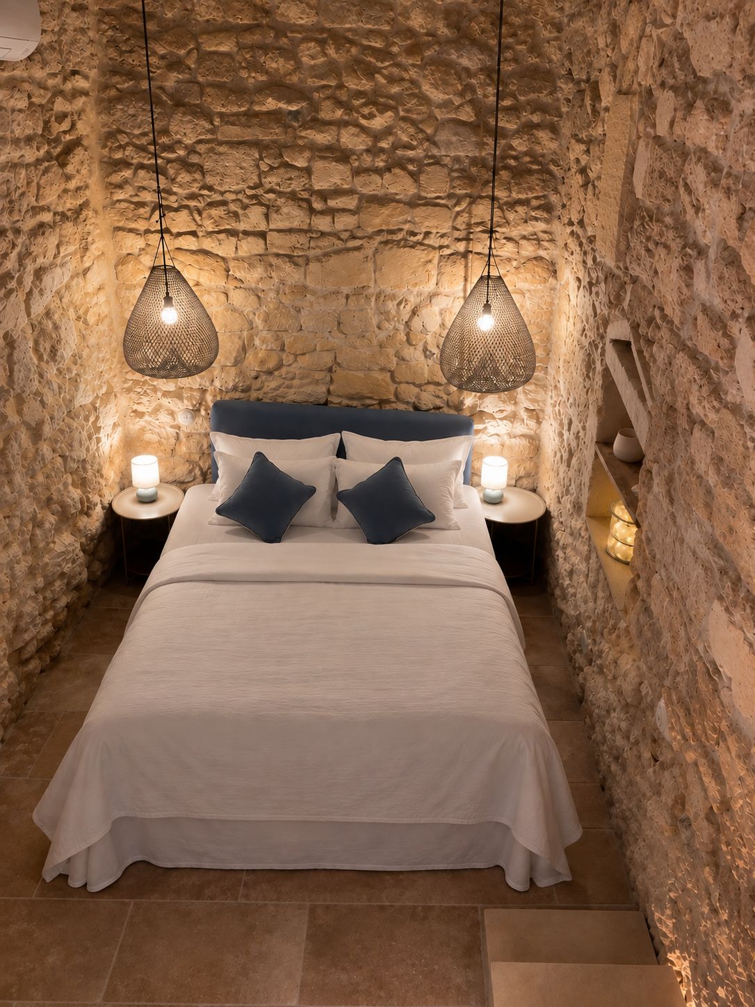 Ein Schlafzimmer mit einer Steinsandsteinwand, einem Bett mit weißer Bettwäsche und dunklen Kissen, zwei Nachttischlampen und einer Pendelleuchte.