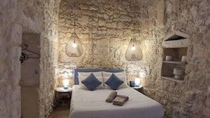 Un dormitorio con paredes de piedra, una cama, dos lámparas colgantes, una mesita y una estantería con adornos.