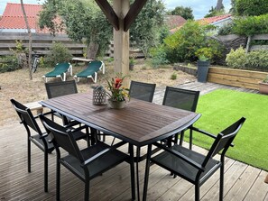 An outdoor wooden deck with a table and chairs, surrounded by greenery and a view of a building with a red roof. - MAISON D'Y (Moliets et Maa)