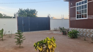 A building with a red exterior and a white window frame, a blue gate, and small plants in the yard. - North Gates - 3 Spacious Ensuite Family Home (Dodoma)