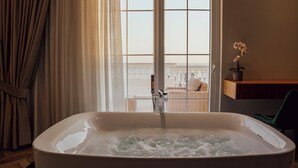 Une salle de bain avec une baignoire, une fenêtre avec des rideaux, un fauteuil en osier et un pot d’orchidée.