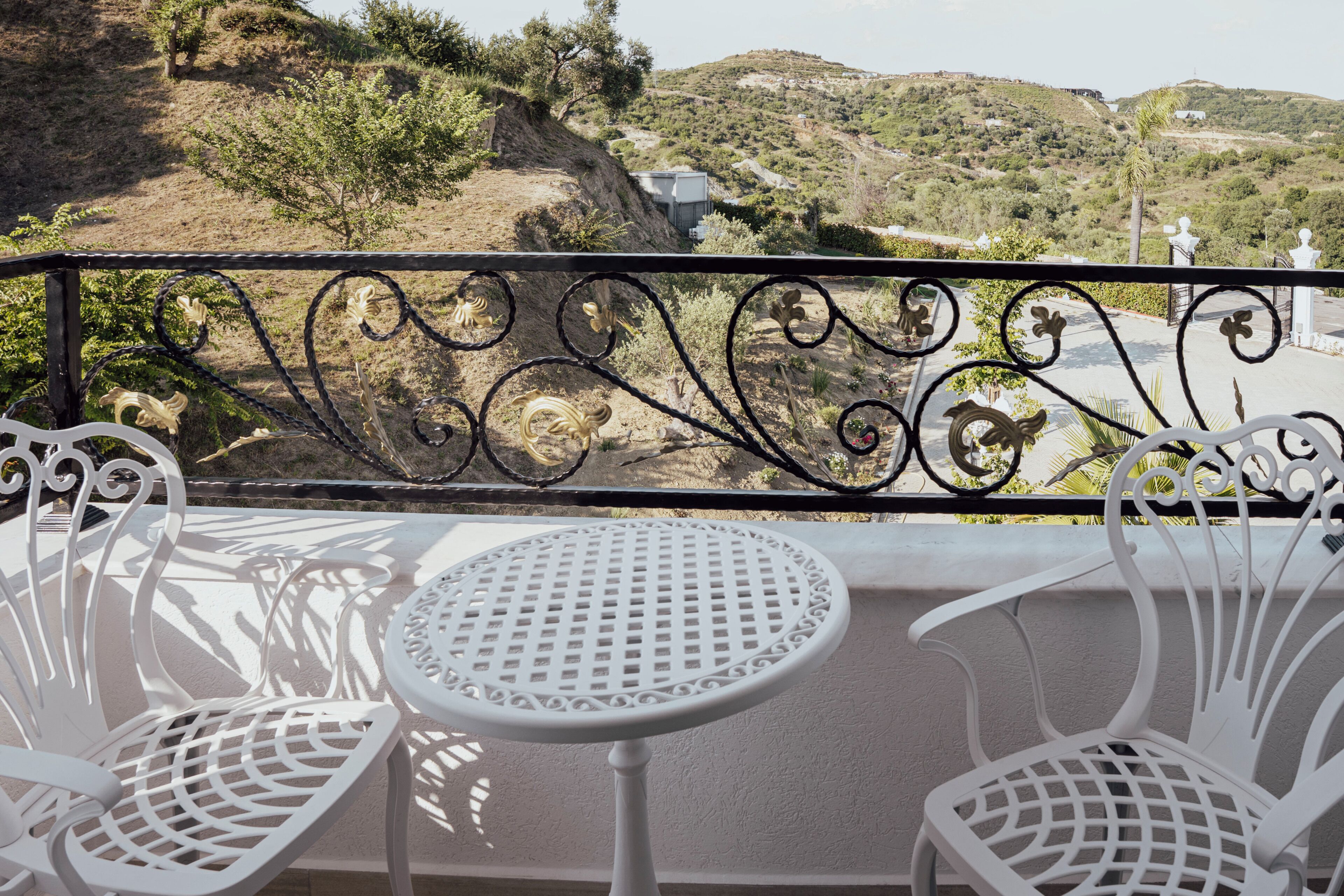 A balcony with white metal chairs and a small round table, overlooking a hilly landscape with sparse vegetation.