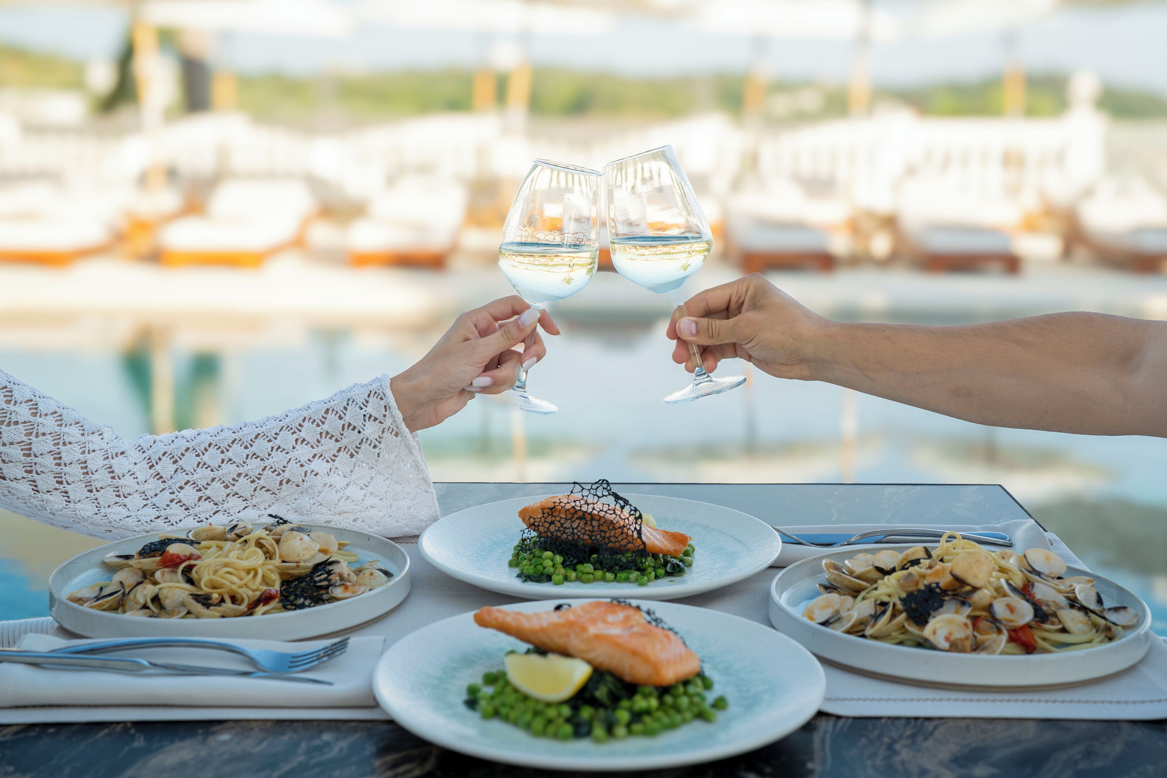 A table with plates of seafood pasta, two people toasting with wine glasses, and a marina view in the background.