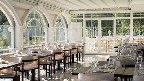 Un restaurant avec un plafond haut, de grandes fenêtres en arc de cercle et une vue sur la verdure environnante. Les tables sont dressées avec des nappes blanches et des verres à vin.