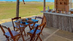 A covered outdoor dining area with a thatched roof, wooden chairs, a table set with food, and a view of the landscape. - Vila Ronquinha (Tutóia)