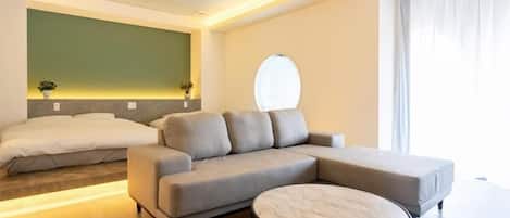A modern hotel room with a sectional sofa, a round coffee table, a bed, and a large window with curtains.