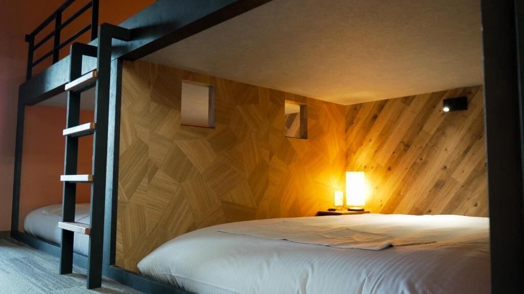 A bunk bed room with a wooden wall, a ladder, a bedside table with a lamp, and a bed with white bedding.