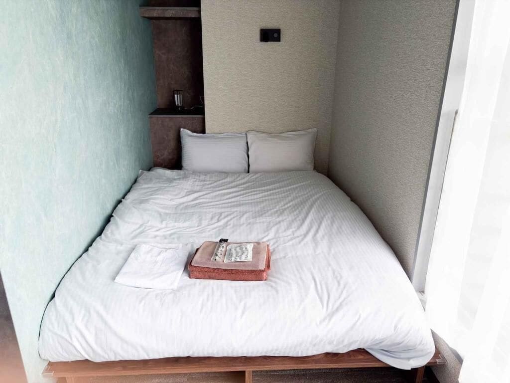 Ein ordentlich bezogenes Bett mit weißer Bettwäsche, ein kleiner Holfnachtisch und eine Wandleuchte.