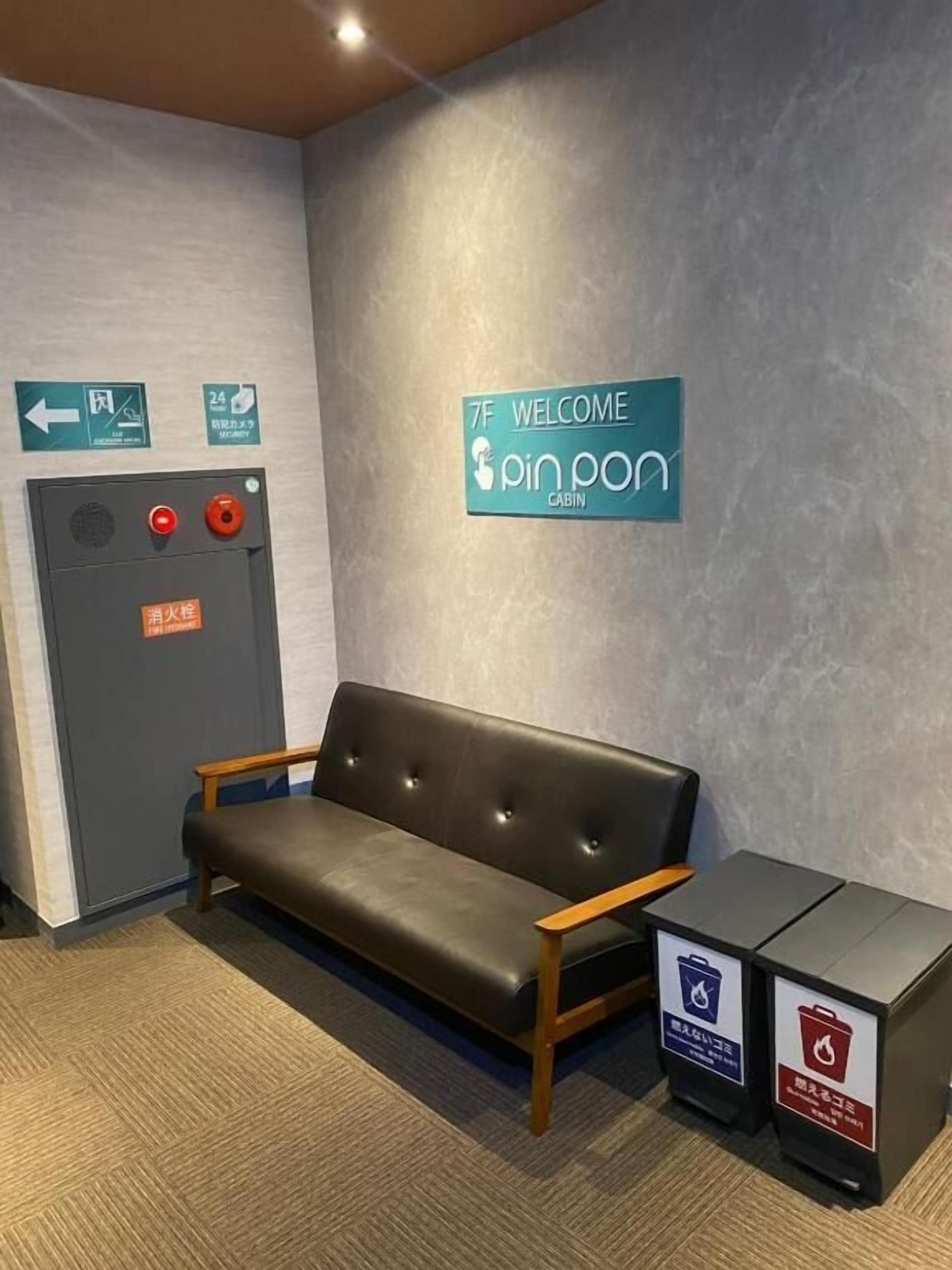 A reception area with a couch, a sign reading '7F Welcome Pinpon Cabin,' a fire alarm panel, and two trash bins.
