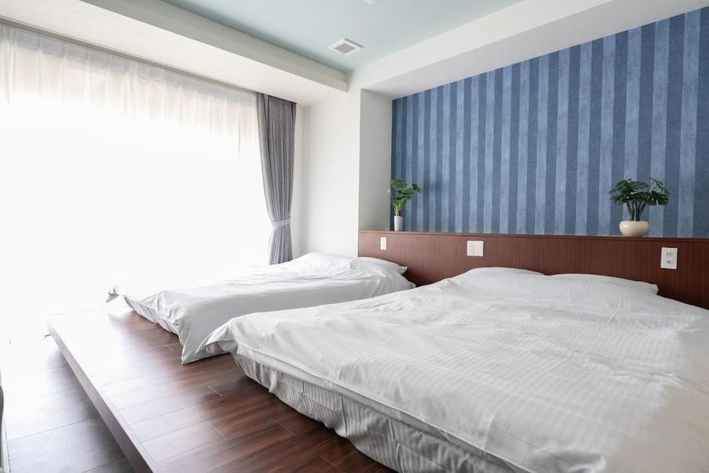 Une chambre d’hôtel avec deux lits, une grande fenêtre avec des rideaux, un mur à rayures bleues et deux plantes en pot.
