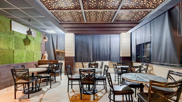 A restaurant interior with a modern design, featuring a patterned ceiling, green wall accents, and a mix of round and square tables with chairs. - Deluxe Room Ranipur Mor (Haridwar)