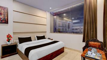 A hotel room with a bed, bedside table, a small table with a tray of breakfast items, a window with curtains, and a painting on the wall.