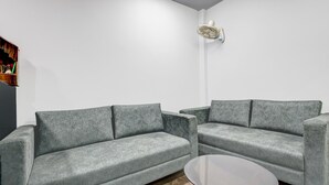 A modern living room with two grey sofas, a small round coffee table, and a wall-mounted fan. - Deluxe Room Ranipur Mor (Haridwar)