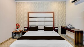 A hotel room with a large bed, a bedside table with a phone, a nightstand, a wall-mounted air conditioner, and a decorative vase with flowers. - Deluxe Room Ranipur Mor (Haridwar)