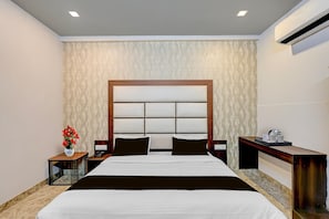 A hotel room with a large bed, a bedside table with a phone, a nightstand, a wall-mounted air conditioner, and a decorative vase with flowers. - Deluxe Room Ranipur Mor (Haridwar)