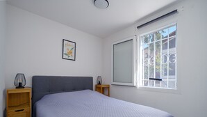 A bedroom with a bed, a nightstand, a drawer unit, a window with bars, and a ceiling light. - House with a garden and a nearby beach (Agde)
