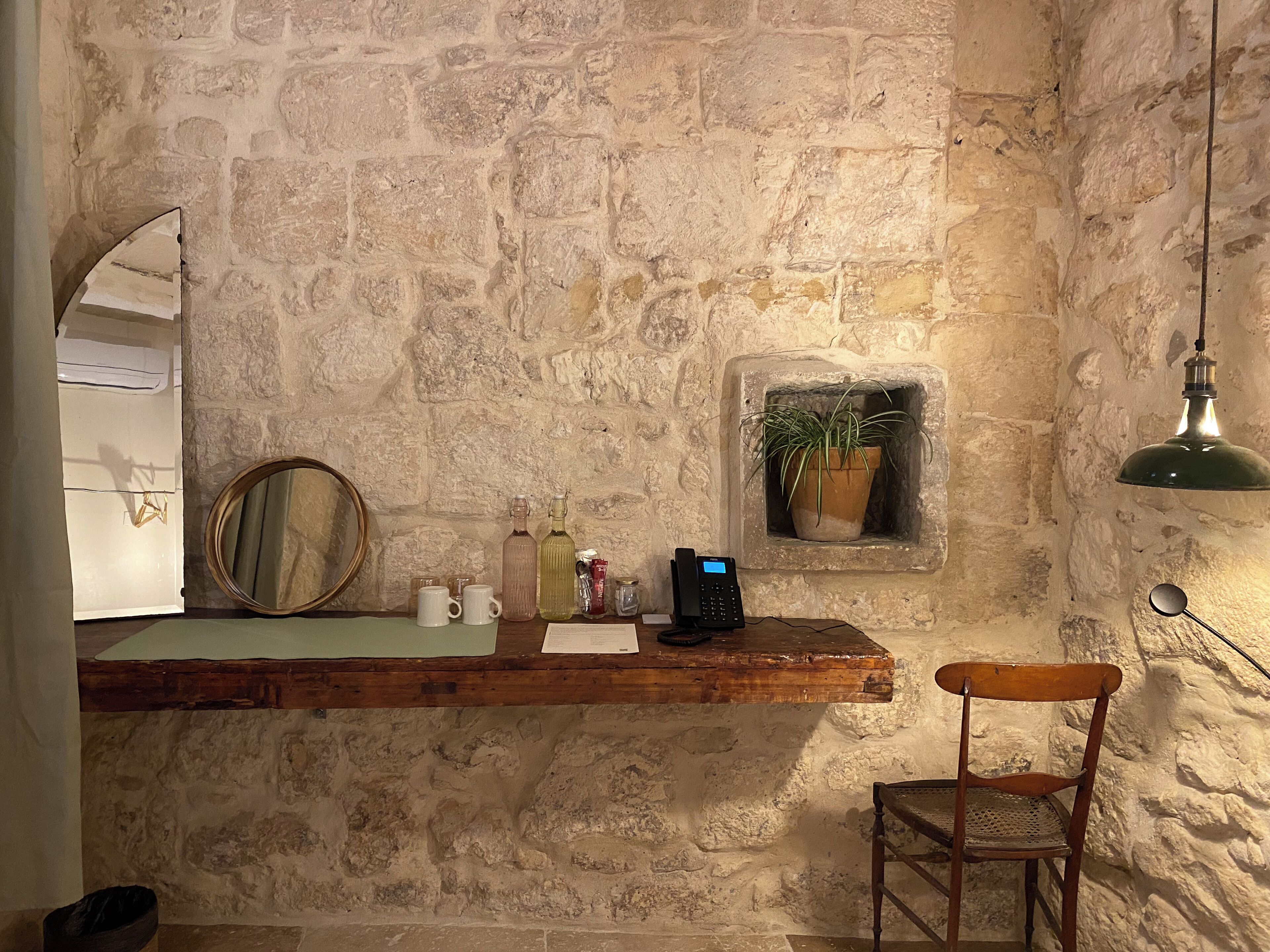 A stone-walled room with a wooden counter, a chair, a mirror, a potted plant, and a hanging light fixture.
