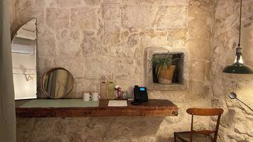 A stone-walled room with a wooden counter, a chair, a mirror, a potted plant, and a hanging light fixture.