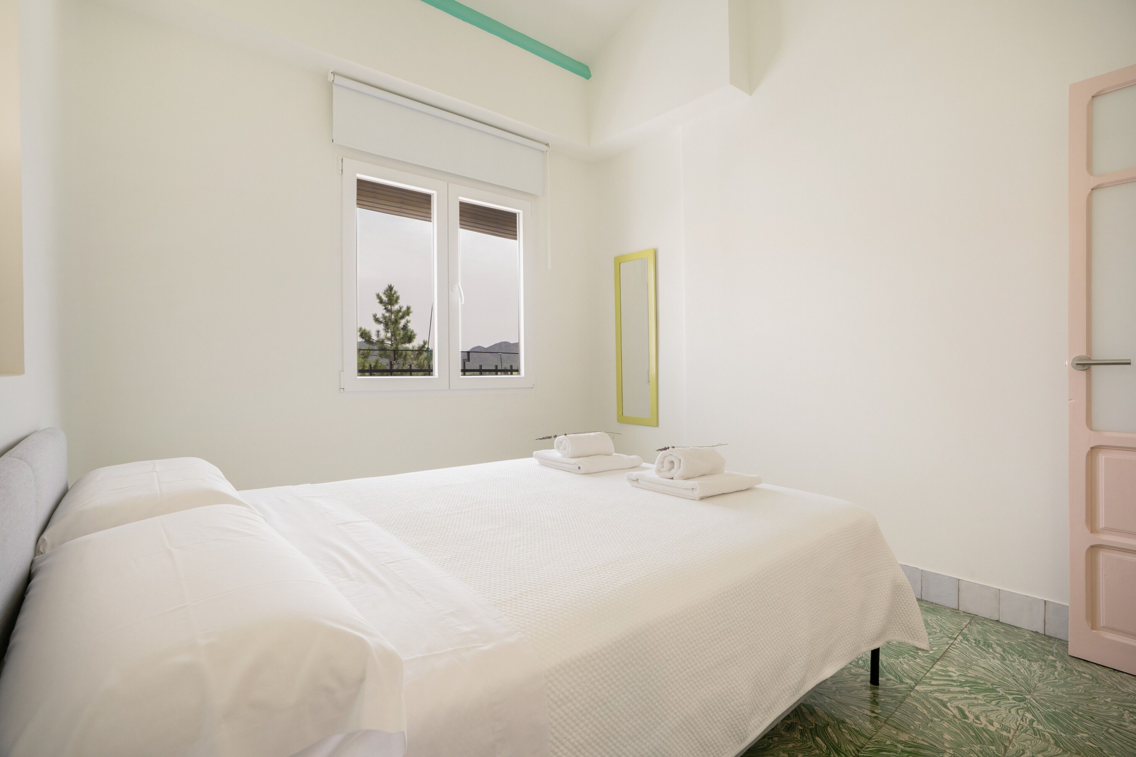 Een hotelkamer met een bed, een raam met zonwering, een spiegel en een deur.