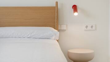 En seng med træsenggavl, et natbord, en vægmonteret lampe og en stikkontakt.