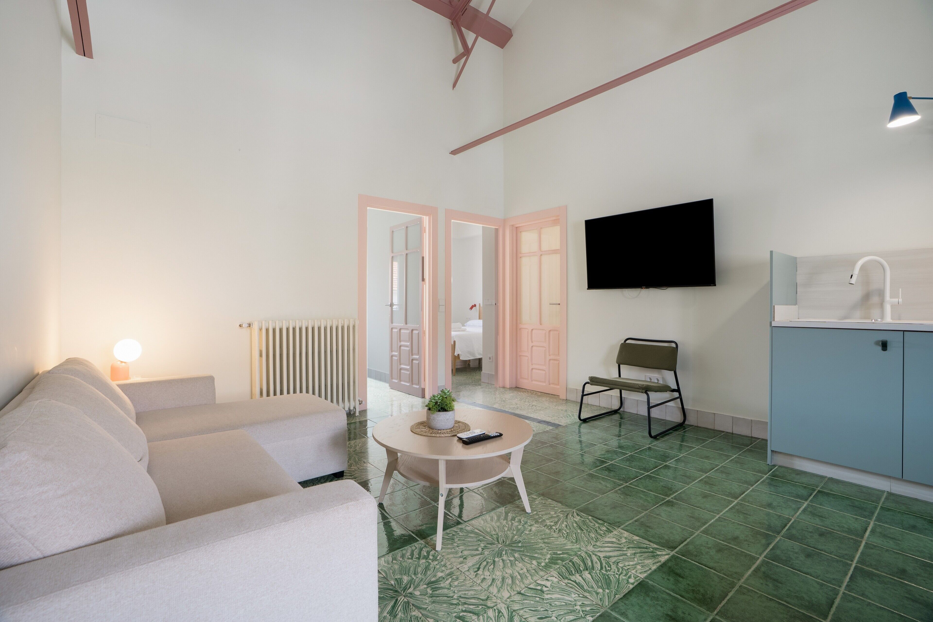 Ein Wohnzimmer mit einer Couch, einem kleinen runden Tisch mit einer Pflanze, einem an der Wand montierten Fernseher und einer Küchenzeile mit Spüle und Schränken.