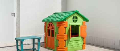 Ein kleines Spielhäuschen mit einem grünen Dach, orangen Wänden, einer blauen Bank und einer weißen Tür im Hintergrund.