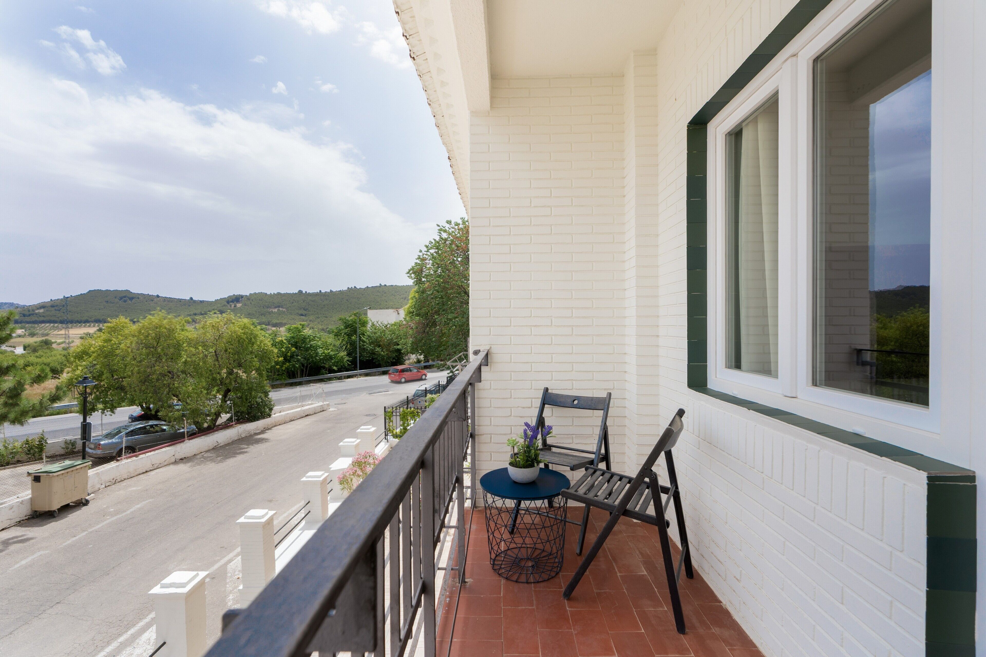 A balcony with a view of a road and hills, a chair, a small table with a plant, and a window with blinds.