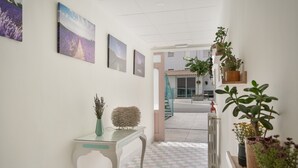 A hallway with a tiled floor, a white wall with framed pictures, a glass-top table with a vase, and potted plants.