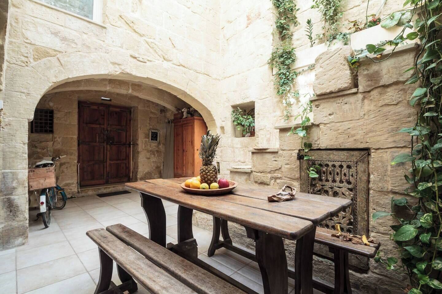 Une cour pavée de pierres, avec une table en bois dressée, sur laquelle on a disposé des fruits, ainsi que des bancs et un vélo.