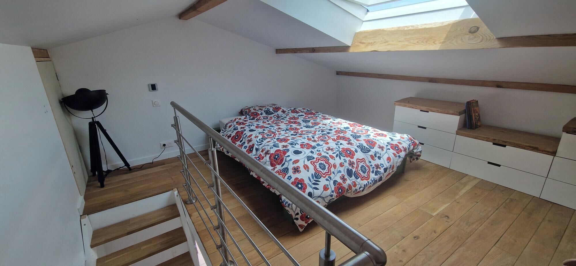 Una camera da letto con soffitto spiovente, un letto con copripiumino floreale, pavimento in legno, una piccola scrivania e un lucernario.
