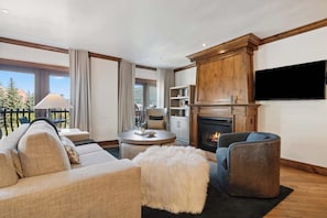 Ein modernes Wohnzimmer mit einer Couch, einem Sessel, einem Hocker, einem Kamin, einem Flachbildfernseher und einem Balkon mit Blick auf Bäume.