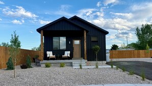 Une maison moderne à l’extérieur sombre, avec un porche à l’avant doté de deux chaises, un petit jardin et un ciel dégagé.