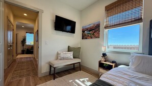 A bedroom with a bed, a TV mounted on the wall, a window with blinds, a painting, and a bedside table with a lamp.