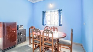 Une pièce avec un mur bleu, une table ronde entourée de chaises, une fenêtre avec des barreaux en métal et un réfrigérateur.
