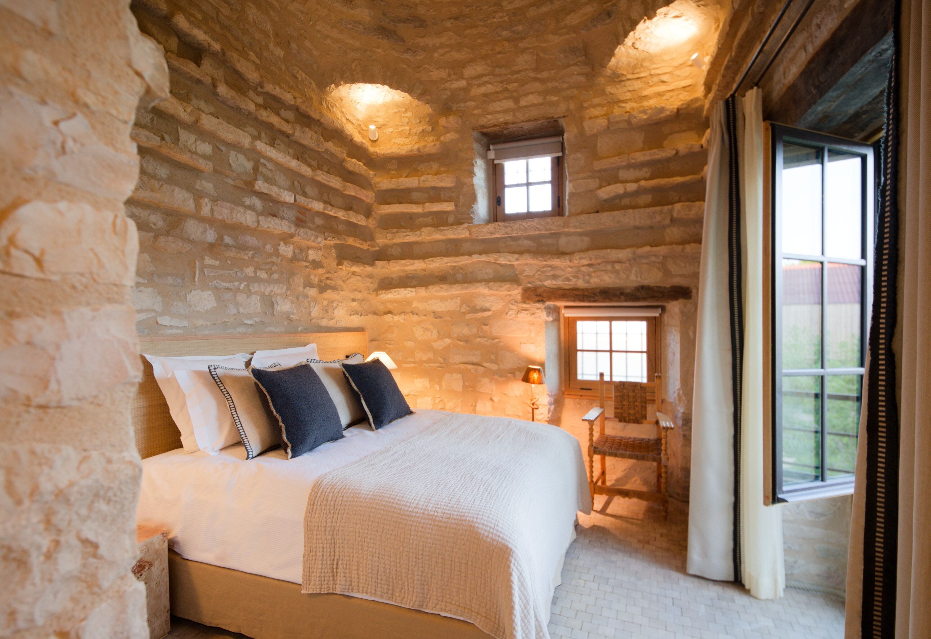 A bedroom with a stone wall, a bed with pillows, a nightstand, a window, and a door.