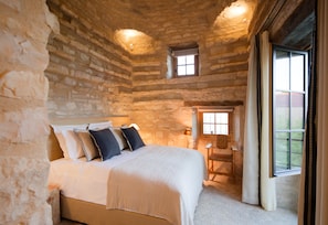 Quarto com parede de pedra, cama com almofadas, mesa de cabeceira, janela e porta.