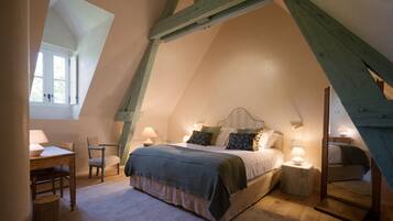 A bedroom with a sloped ceiling, a bed with multiple pillows, a nightstand with a lamp, a chair, and a small table with a lamp.