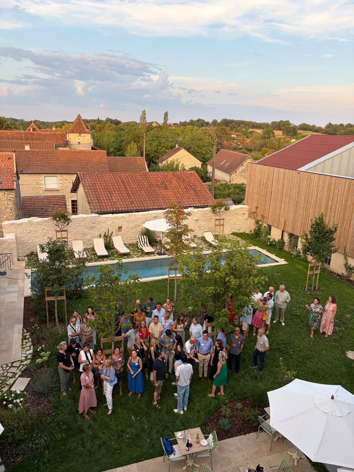Un groupe de personnes s’est réuni dans une cour arrière dotée d’une piscine, entourée de bâtiments en pierre et d’une structure en bois.