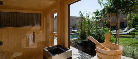 Un sauna de madera con sauna, un balde de madera y vistas a una piscina y follaje exterior.