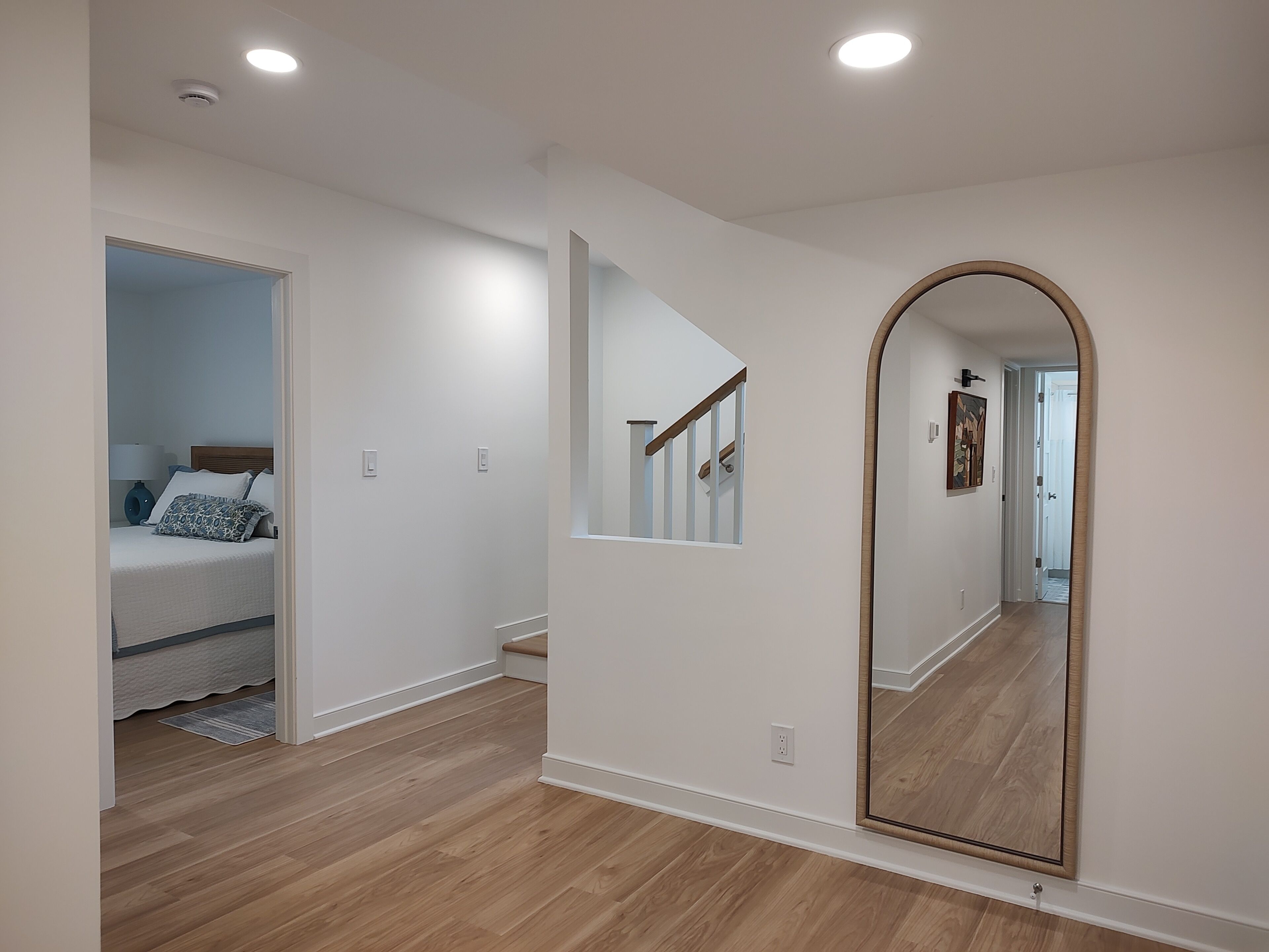 A room with a wooden floor, a bed with a headboard, a large mirror, and a staircase.