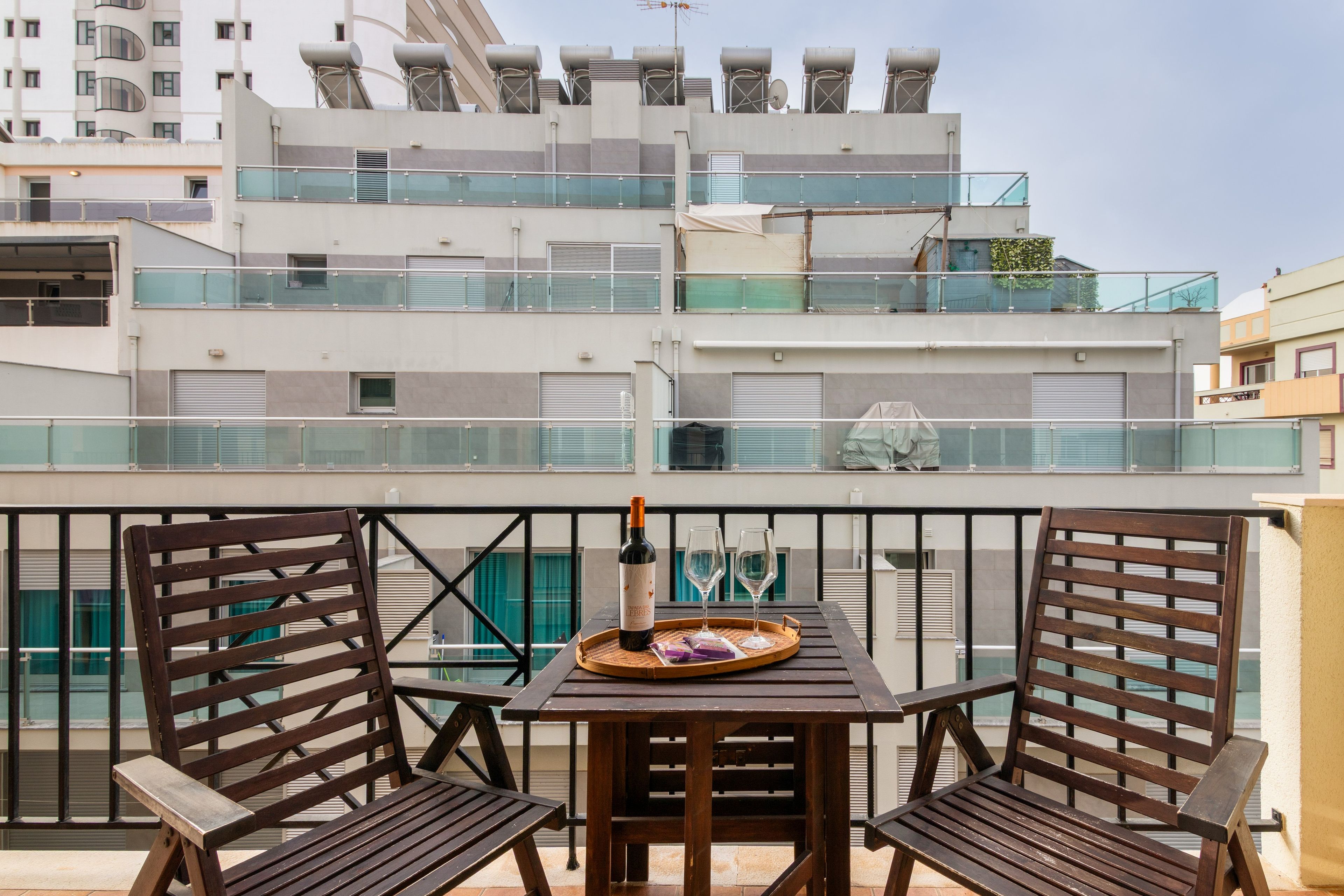 Une terrasse sur le toit, avec une table, deux chaises, une bouteille de vin et un plateau contenant des verres et un sac.